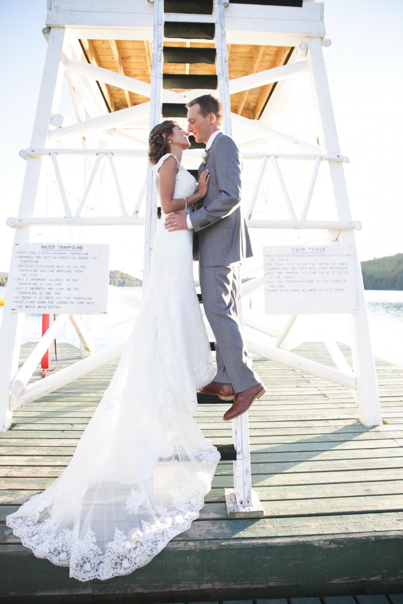 Bride and groom standing on the ladder of a white swim tower at the end of a wooden dock