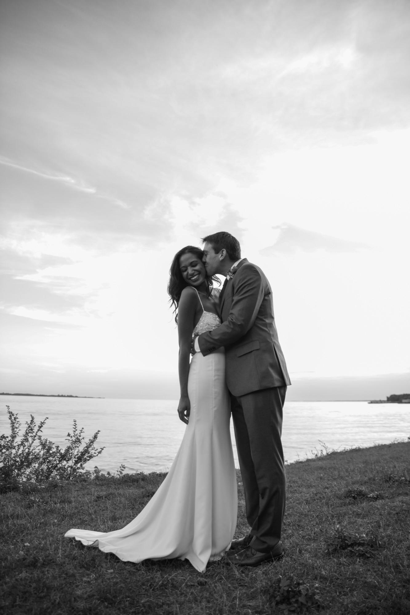 Black and white portrait of a bride and groom on a shoreline under a wide evening sky