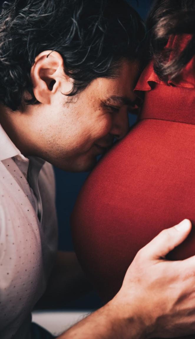 Man with his eyes closed pressing his face gently against his pregnant partner's red dress