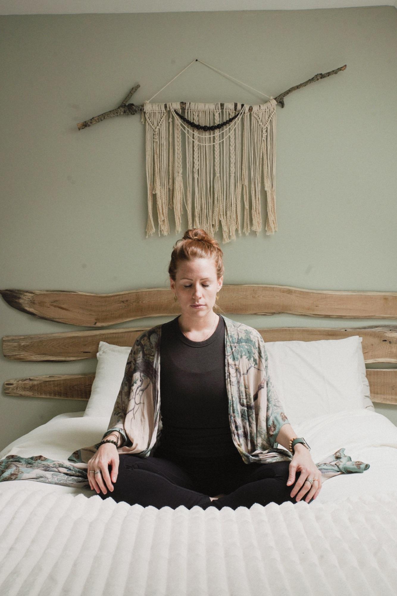 Guest sitting cross-legged and still on a made bed beneath a macrame wall hanging