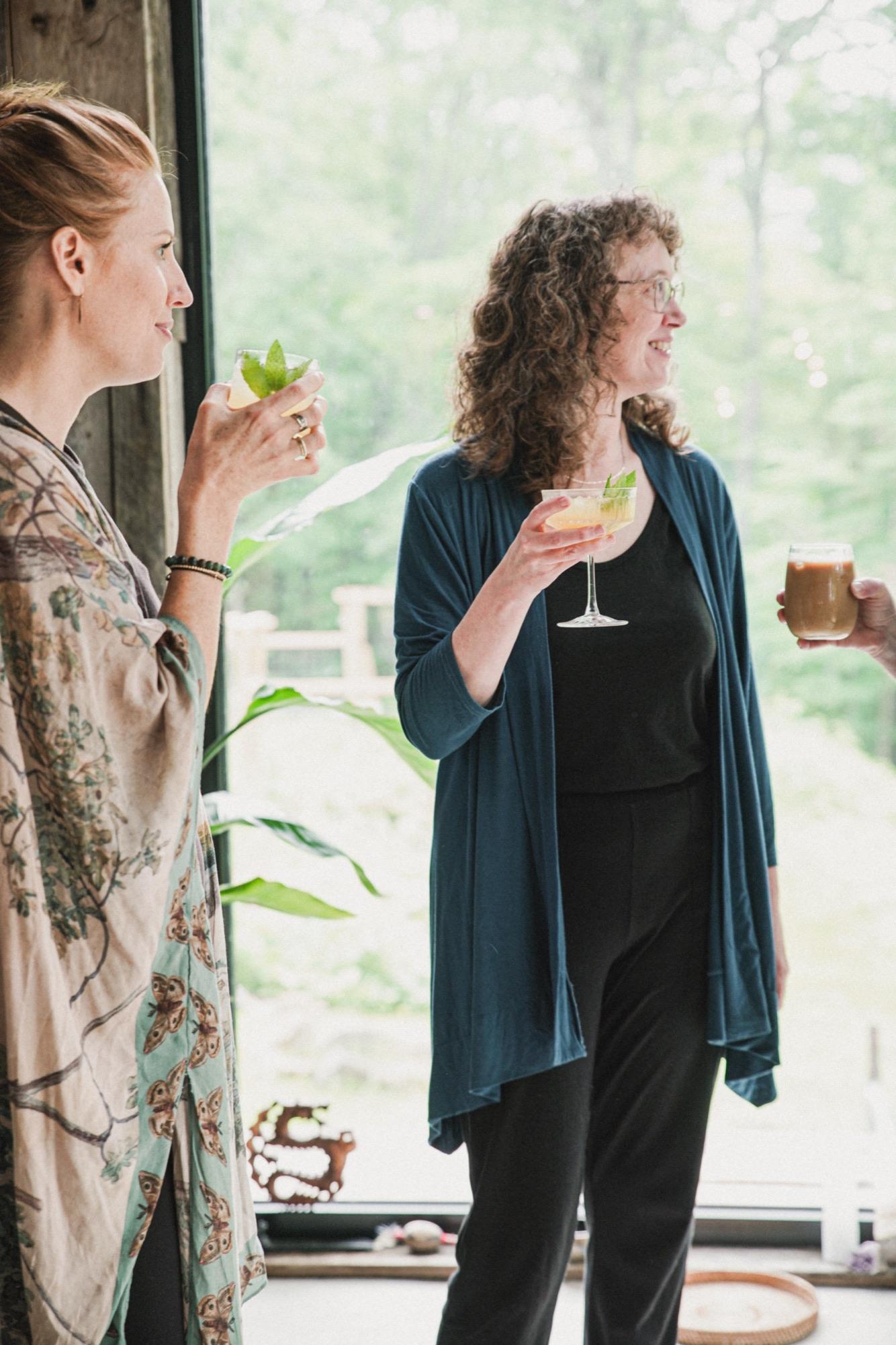 Two guests standing with drinks beside a window looking into green forest