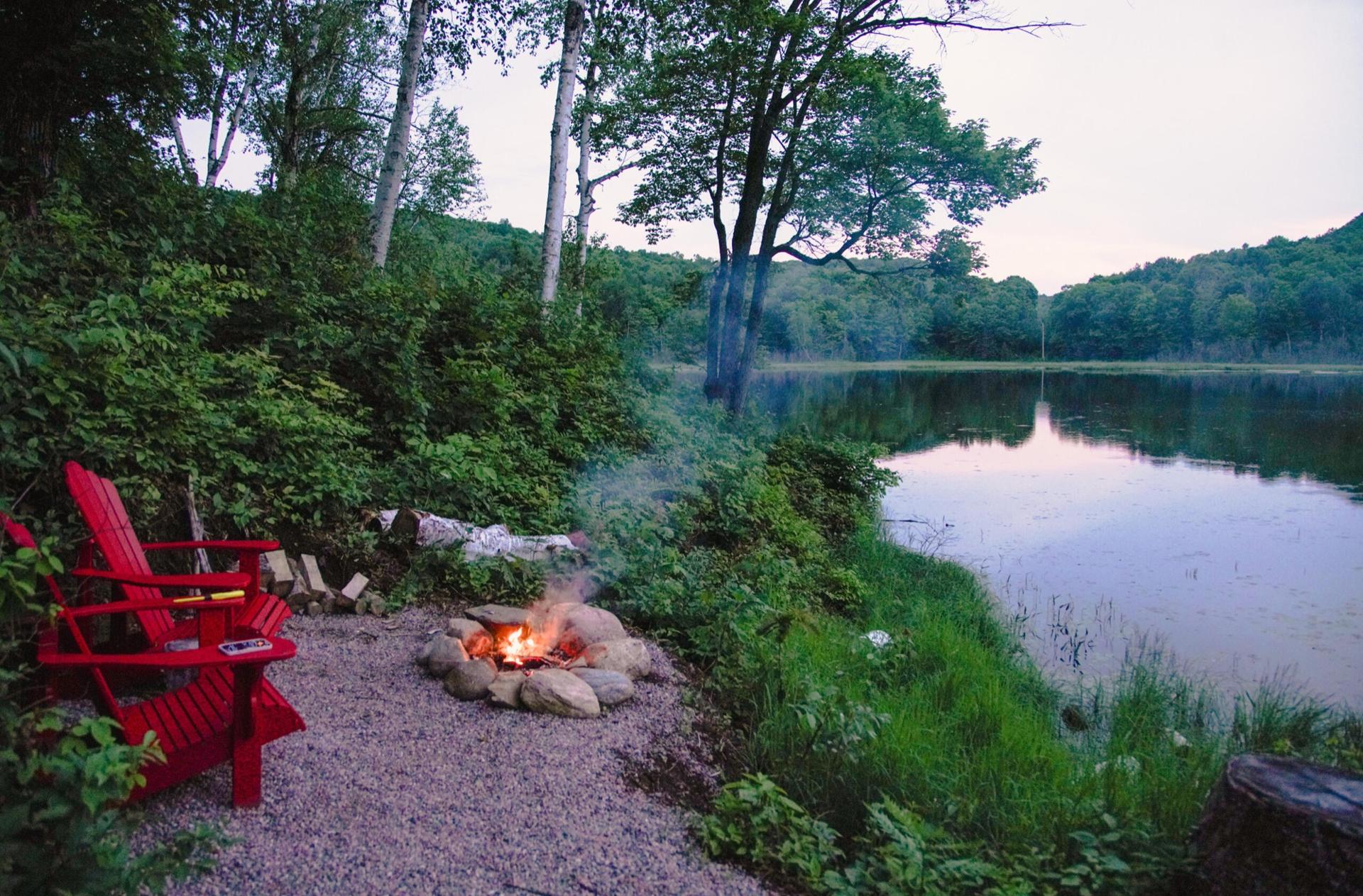 Two red Muskoka chairs beside a lit fire pit at the edge of a still lake at dusk