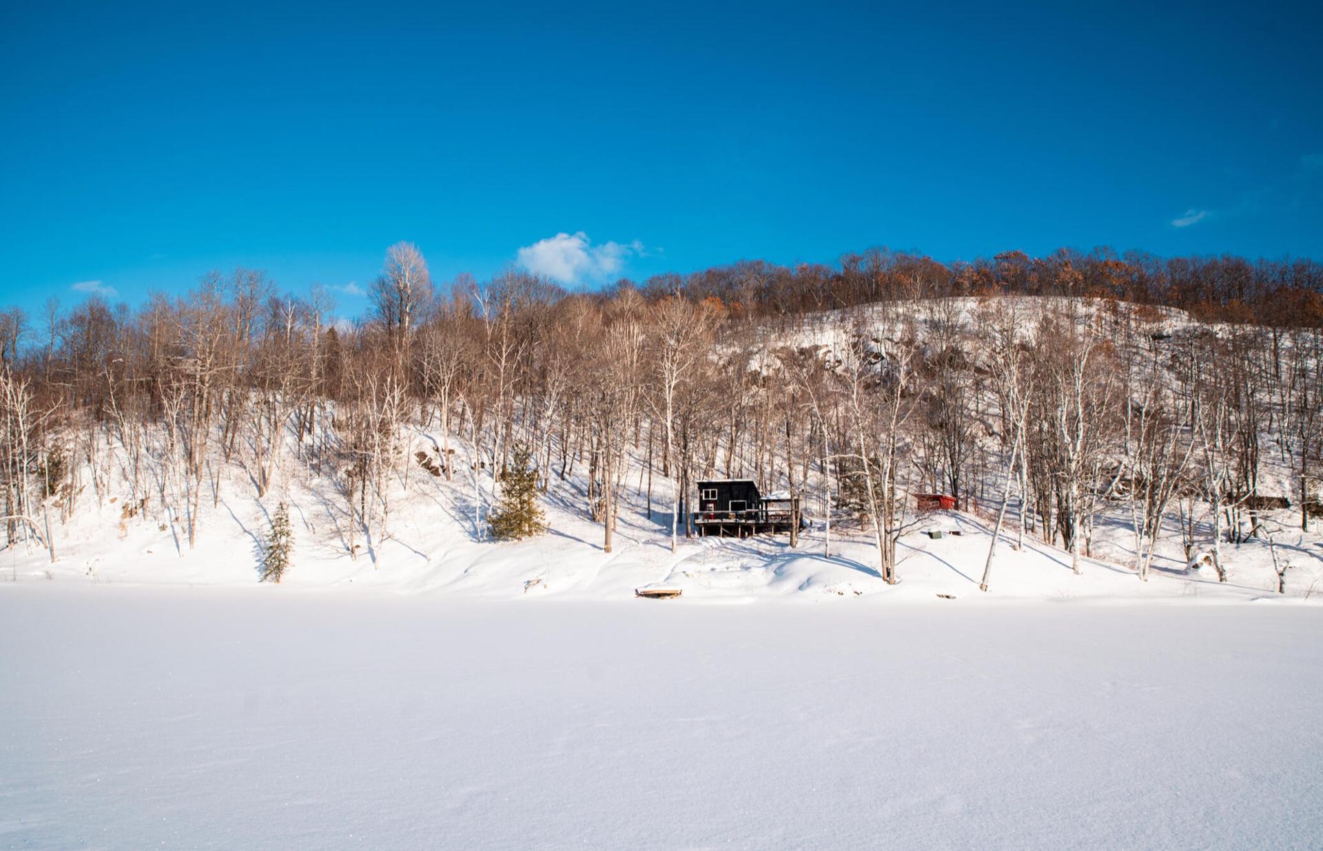 The dark cabin seen from across a snow-covered frozen lake under a clear blue sky
