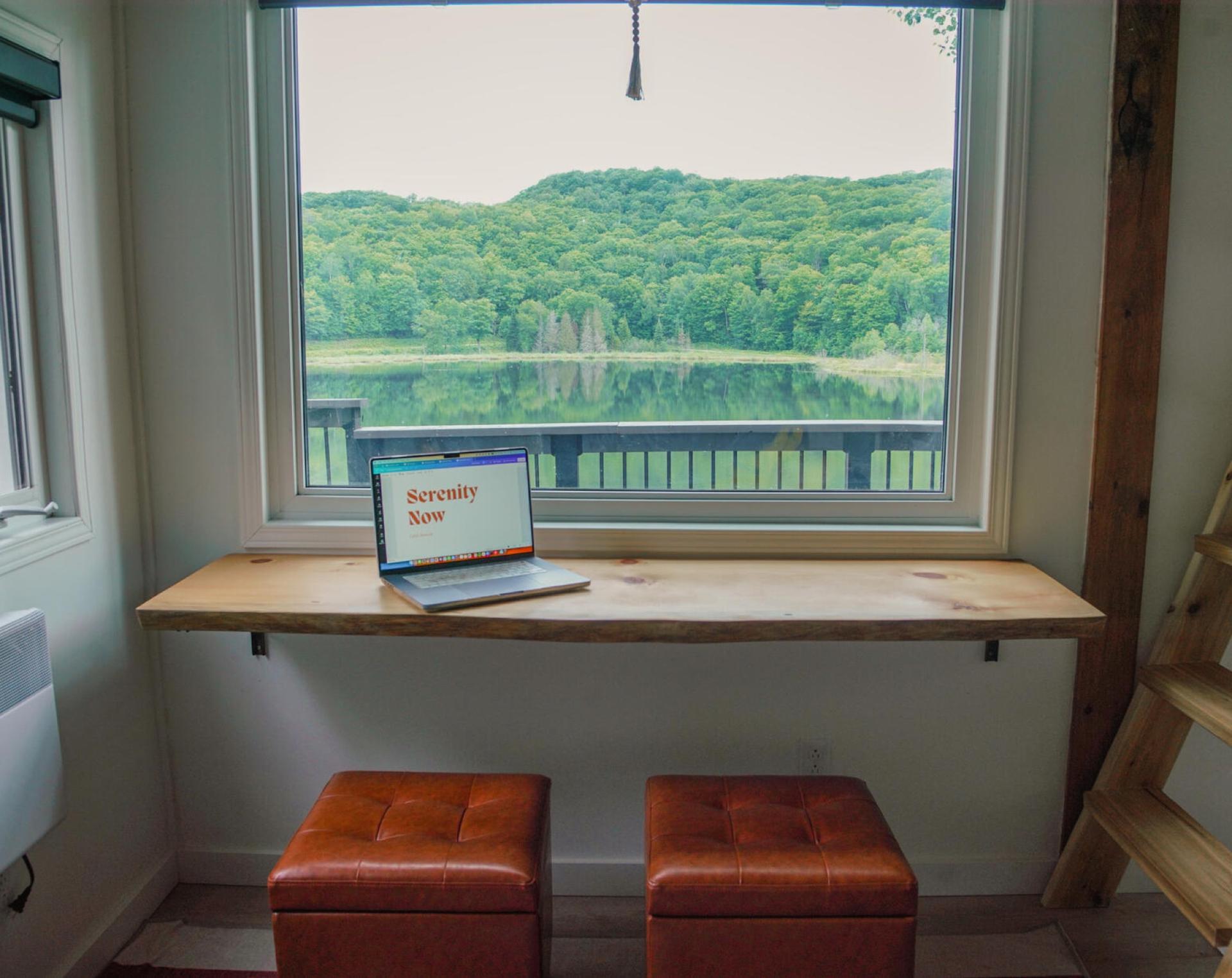 Live-edge desk with a laptop under a wide window looking across the lake to a forested hill