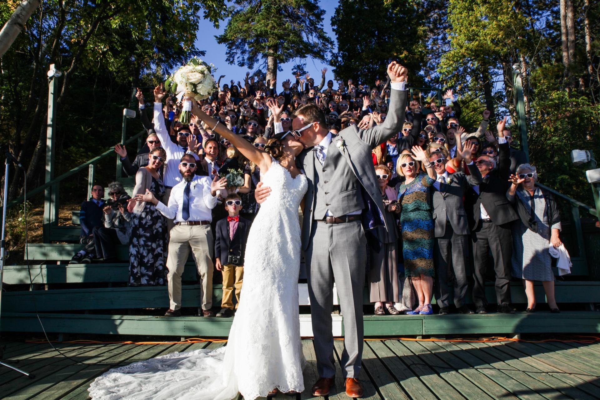 Newly married couple kissing on a lakeside dock as guests in sunglasses cheer from tiered steps behind them