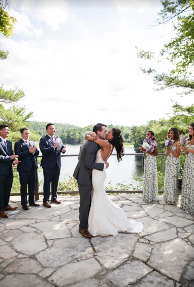 Bride and groom kissing on a flagstone terrace above a lake while the wedding party applauds on either side