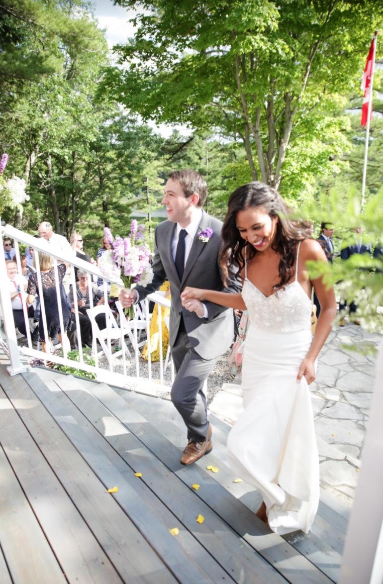 Bride and groom climbing deck steps hand in hand after the ceremony, guests seated behind them and a Canadian flag overhead