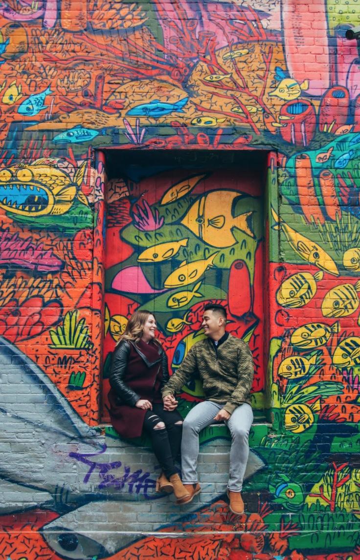 Couple sitting on a ledge in front of a bright mural of orange and yellow fish