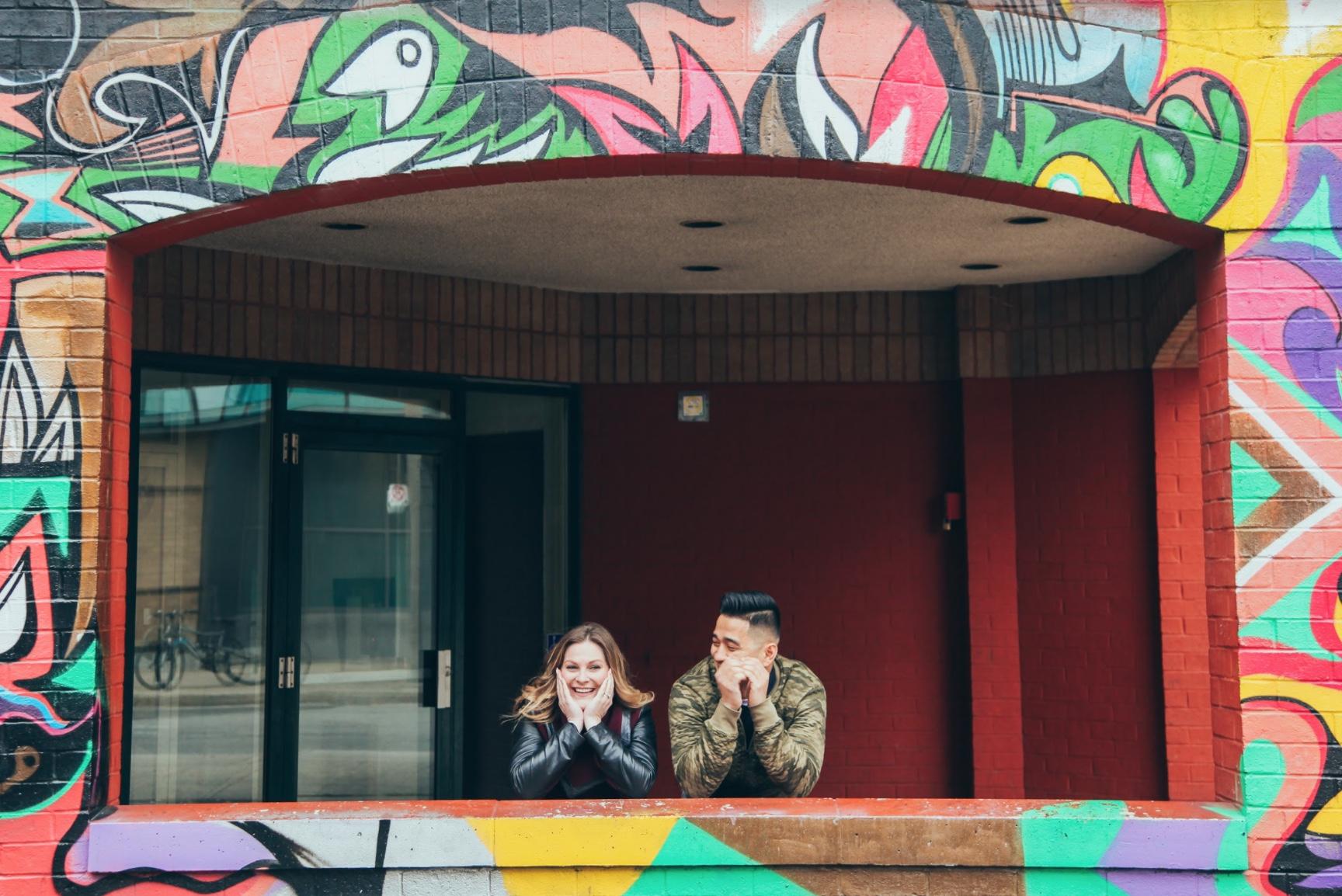 Couple leaning on a painted balcony ledge with their hands on their cheeks, in a building covered in murals