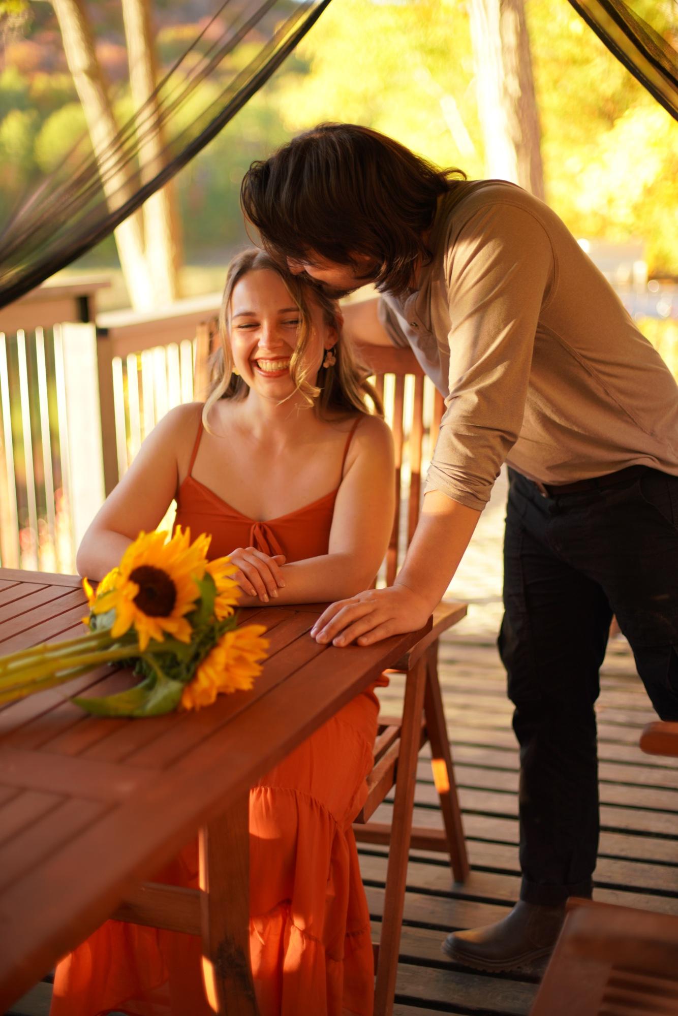 Couple on a cottage deck at golden hour, one leaning in to kiss the other, sunflowers on the table