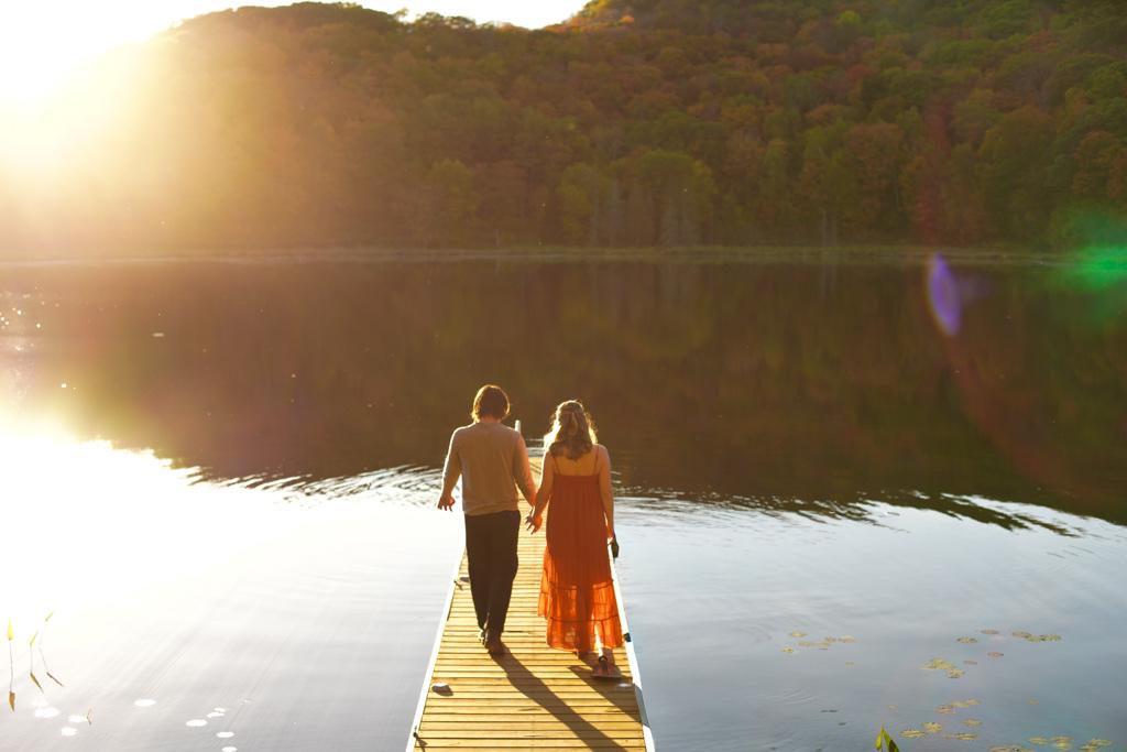 Couple walking hand in hand to the end of a dock at sunset, autumn hills across the lake