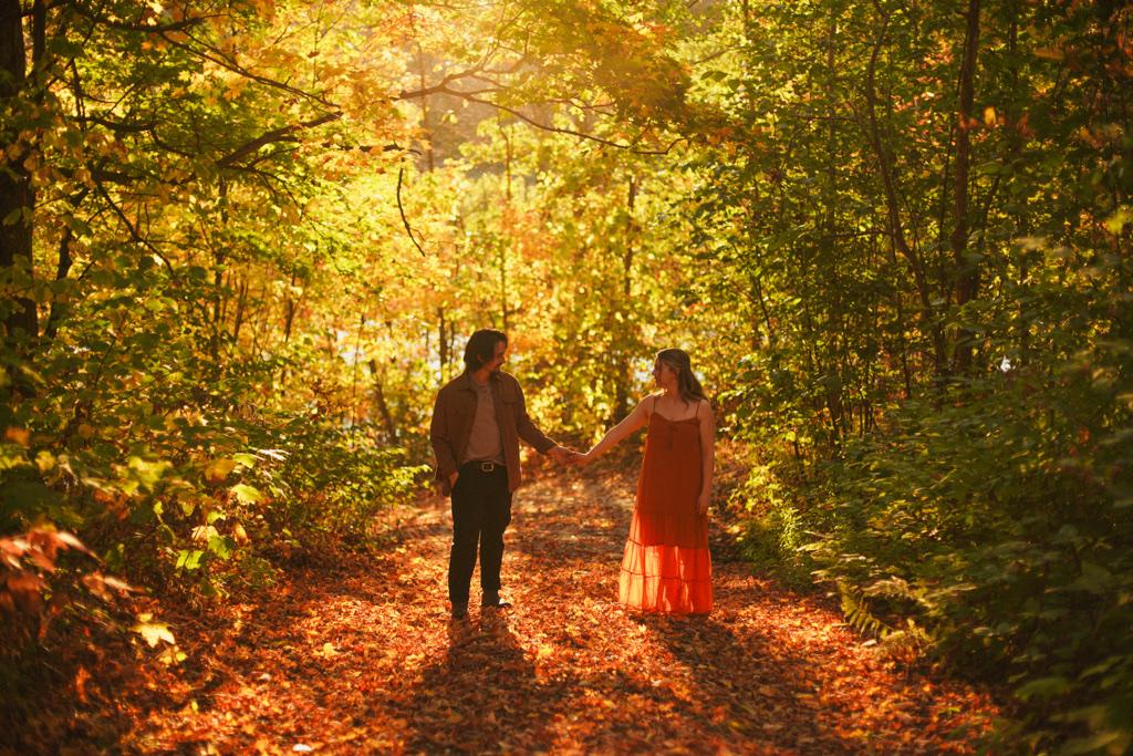 Couple holding hands on a sunlit forest trail carpeted with fallen leaves