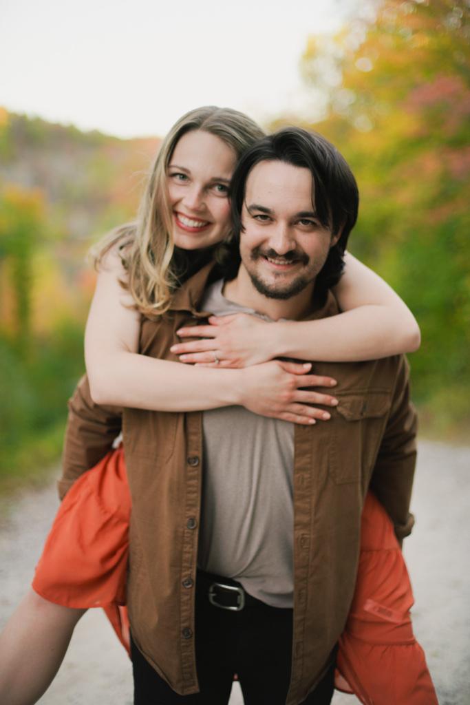 Woman riding piggyback on her partner, both smiling, fall colour behind them