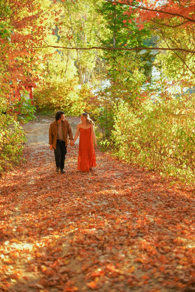 Couple walking hand in hand along a leaf-covered trail under red and orange maples