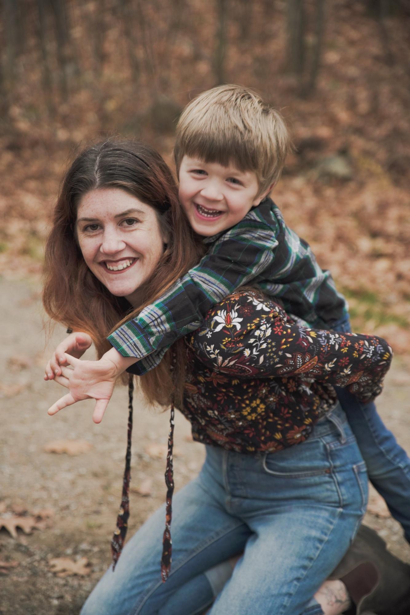 Laughing mother with her young son riding on her back on a leaf-covered autumn trail