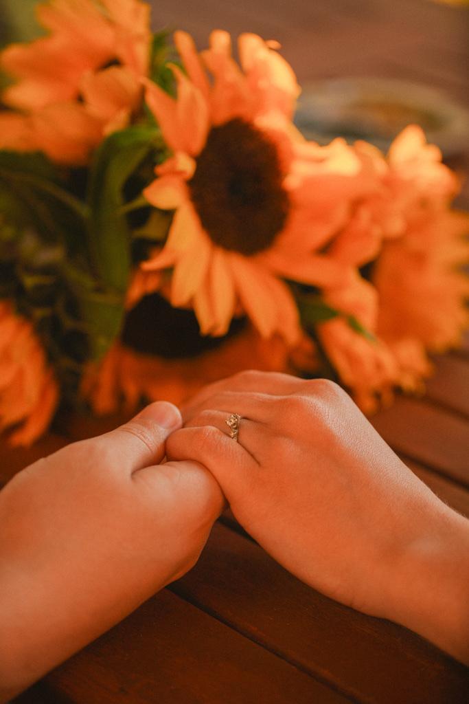 Close detail of a couple's clasped hands and engagement ring beside sunflowers on a wooden table