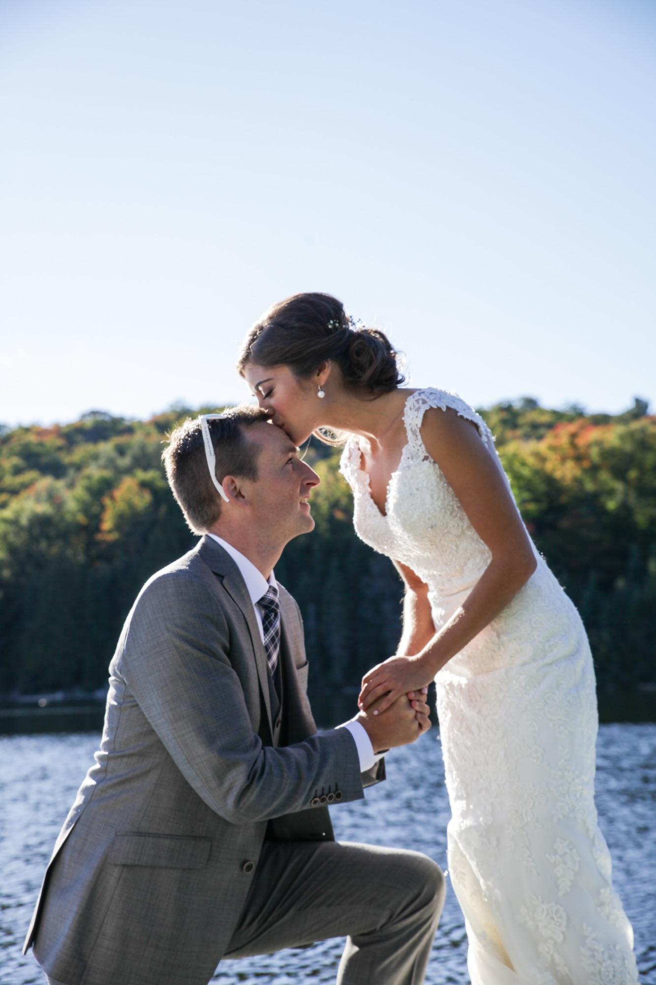 Groom kneeling on a dock while the bride leans down to kiss his forehead, autumn hills across the lake behind them