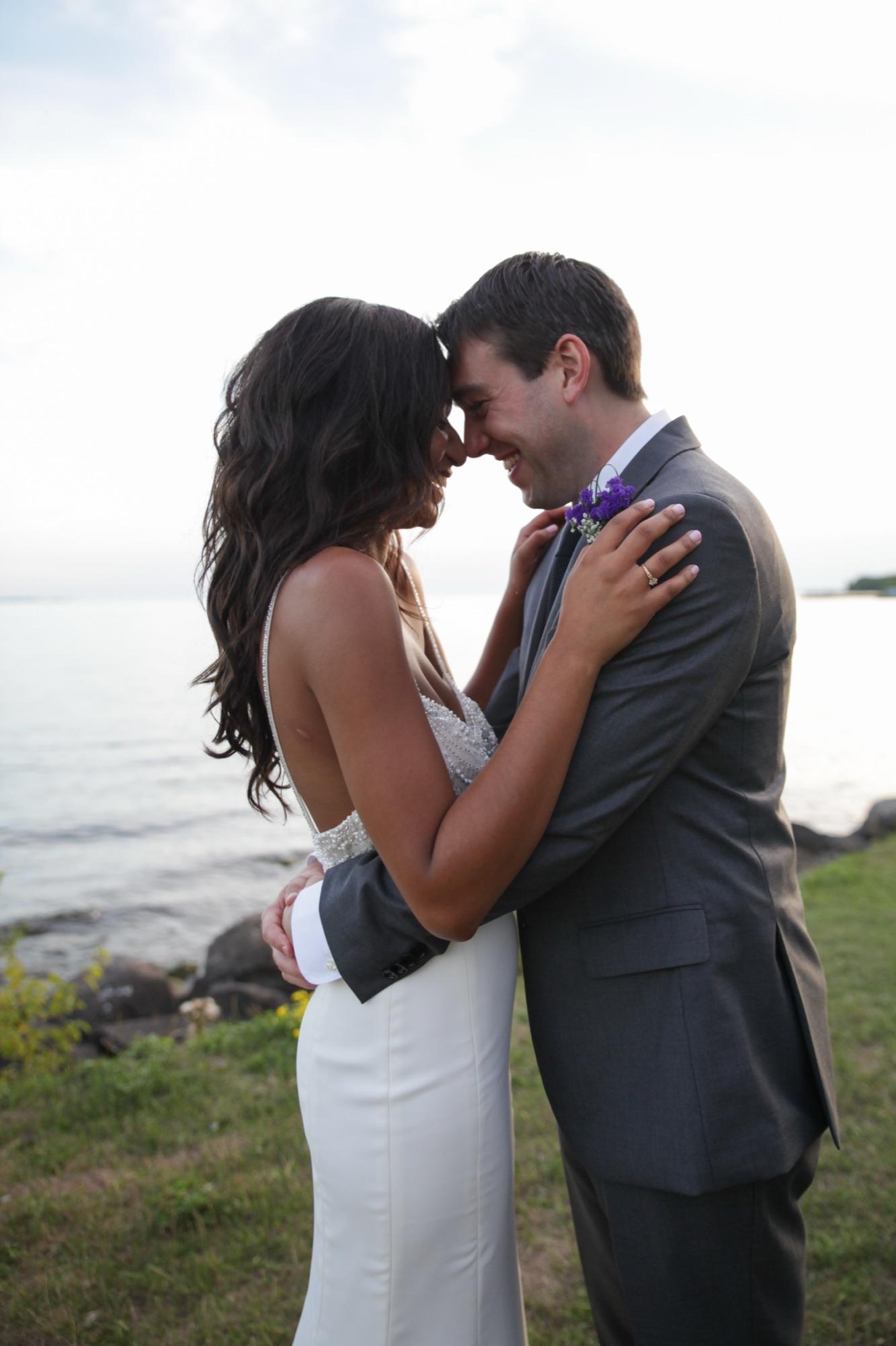 Bride and groom laughing forehead to forehead on a grassy shoreline at the end of the day