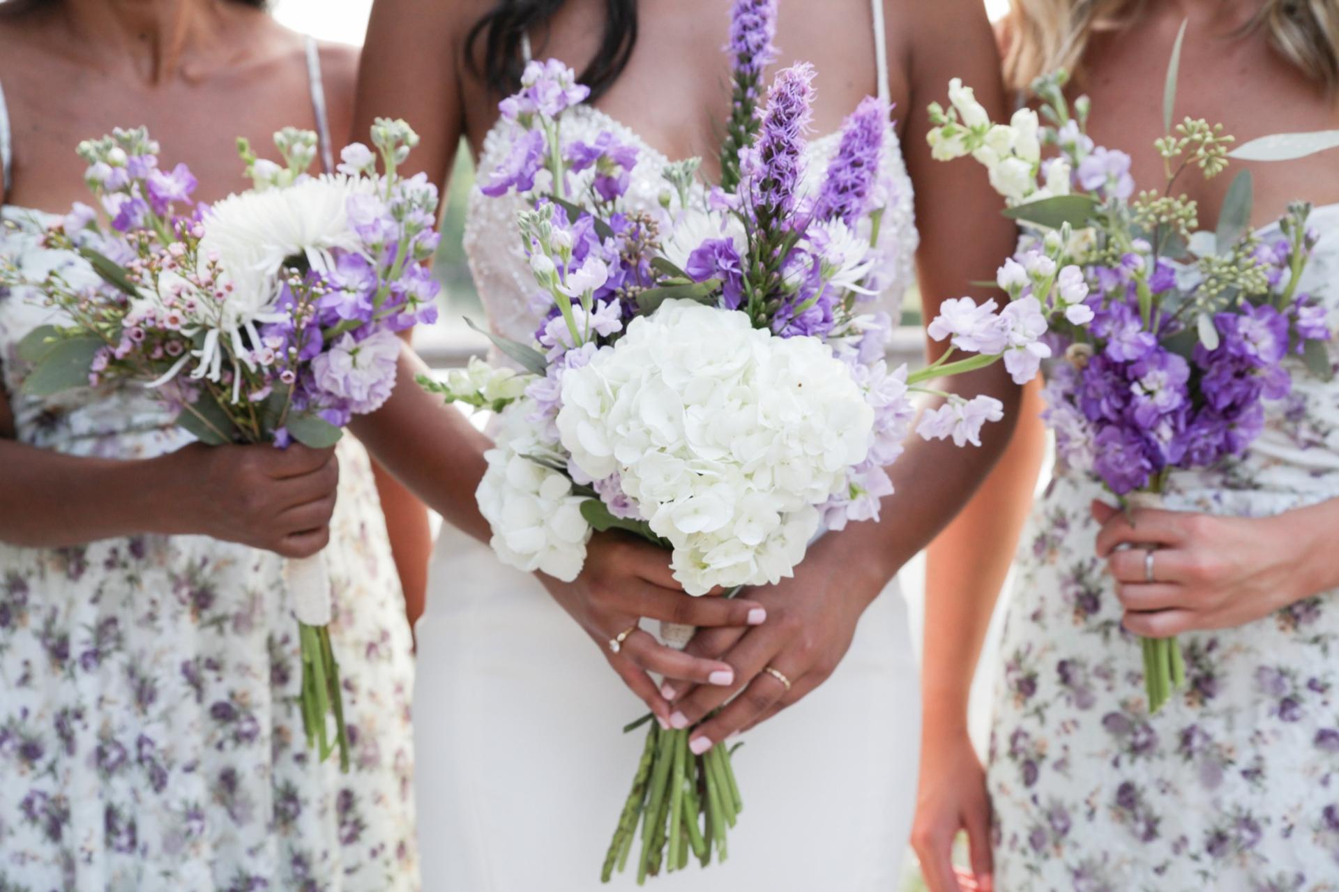 Bride and two bridesmaids holding white and purple bouquets, photographed close in at waist height