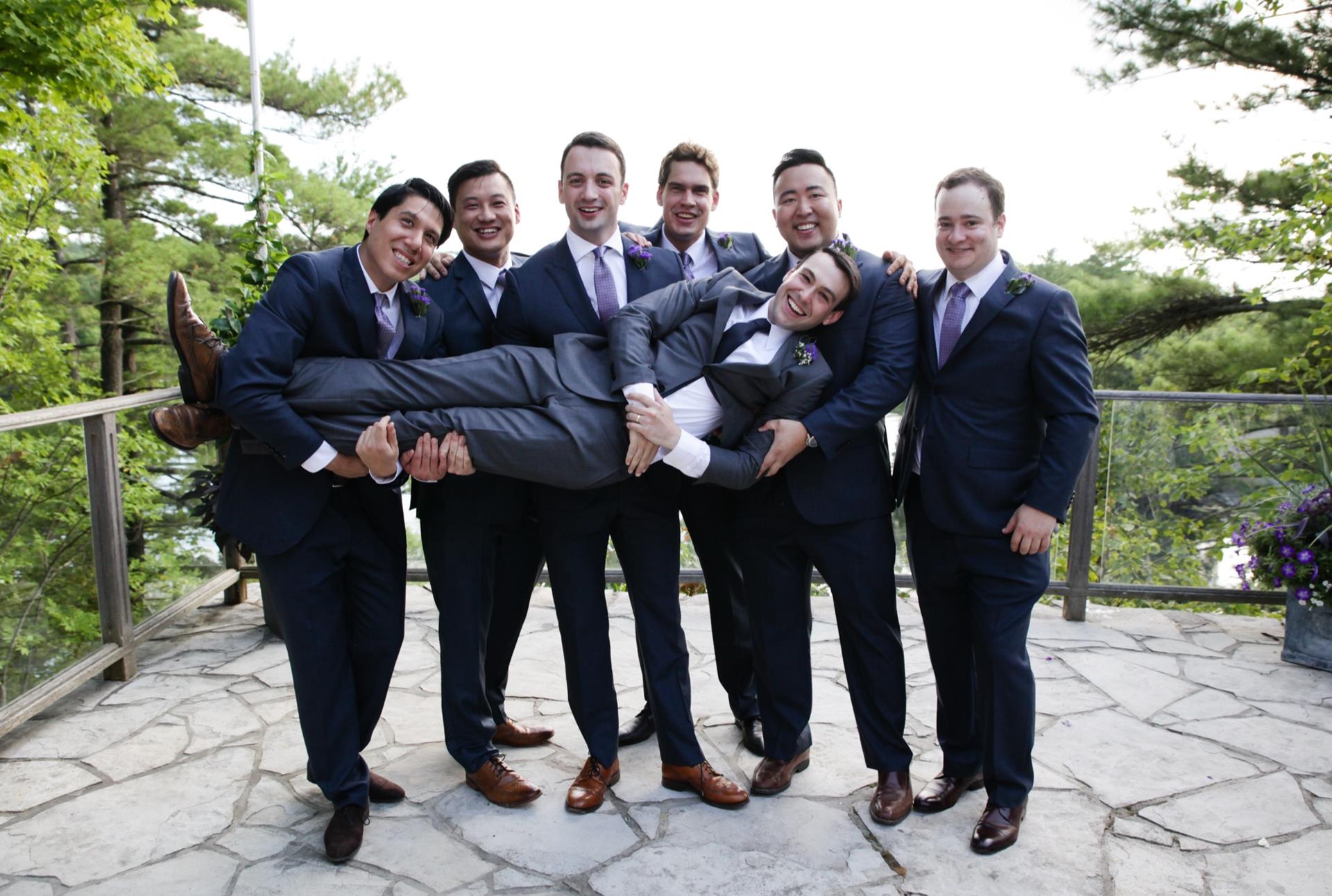 Groomsmen in navy suits carrying the groom horizontally on a flagstone terrace above the water
