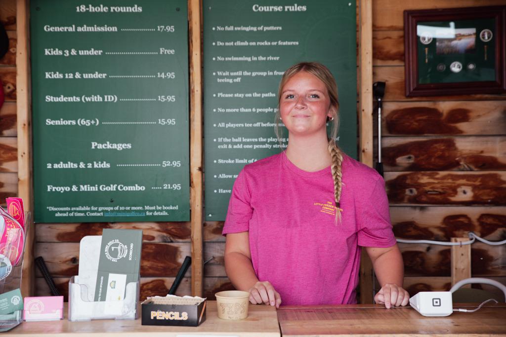 A staff member at the shop counter in front of the price board and course rules