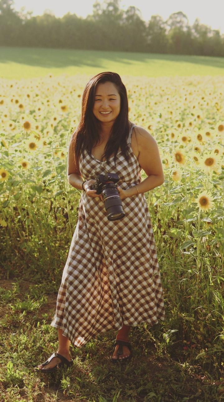 Rosa Park smiling in a sunflower field in a brown gingham dress, holding her camera