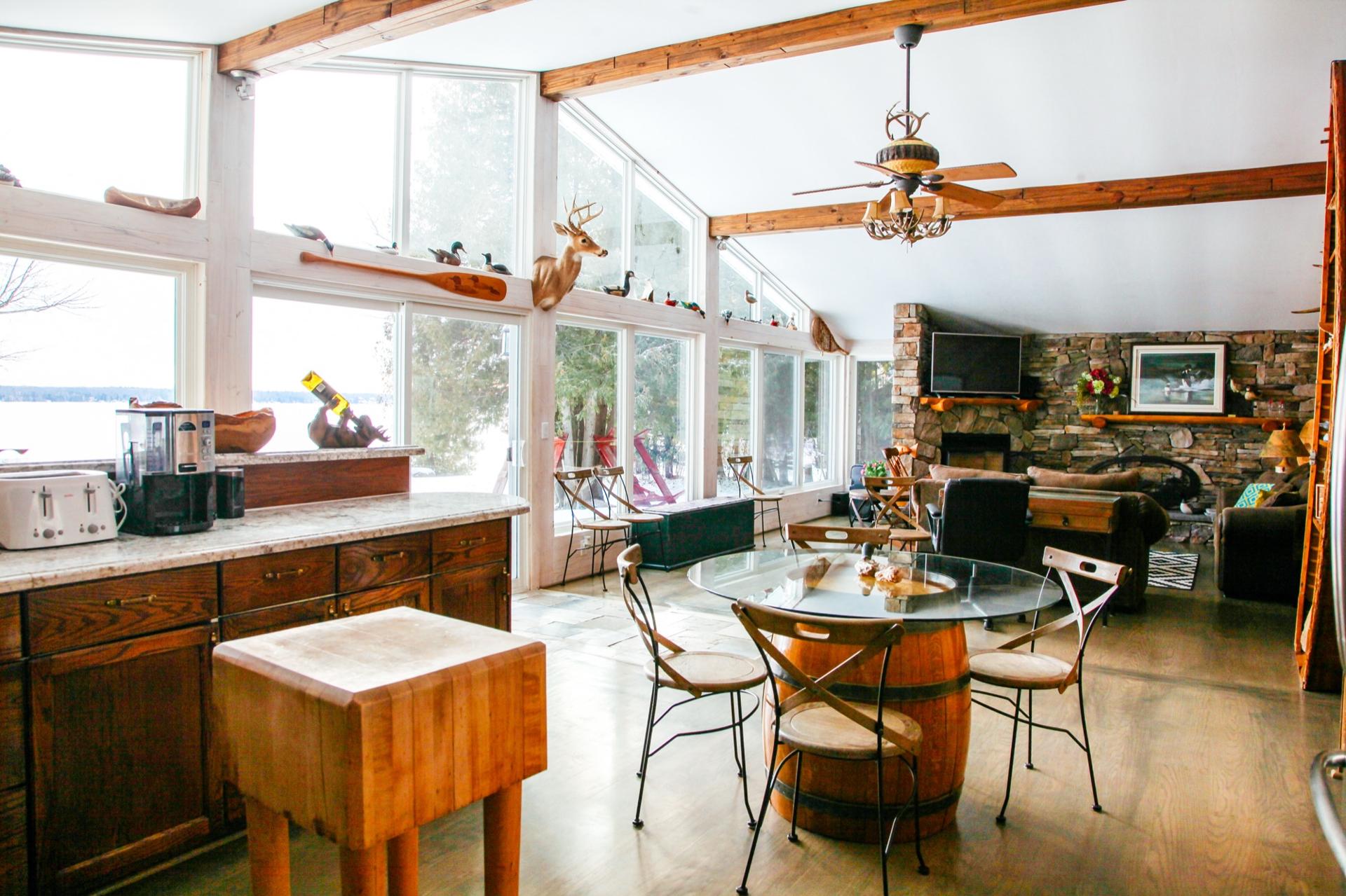Open cottage great room with a stone fireplace, exposed beams and a wall of windows facing the water