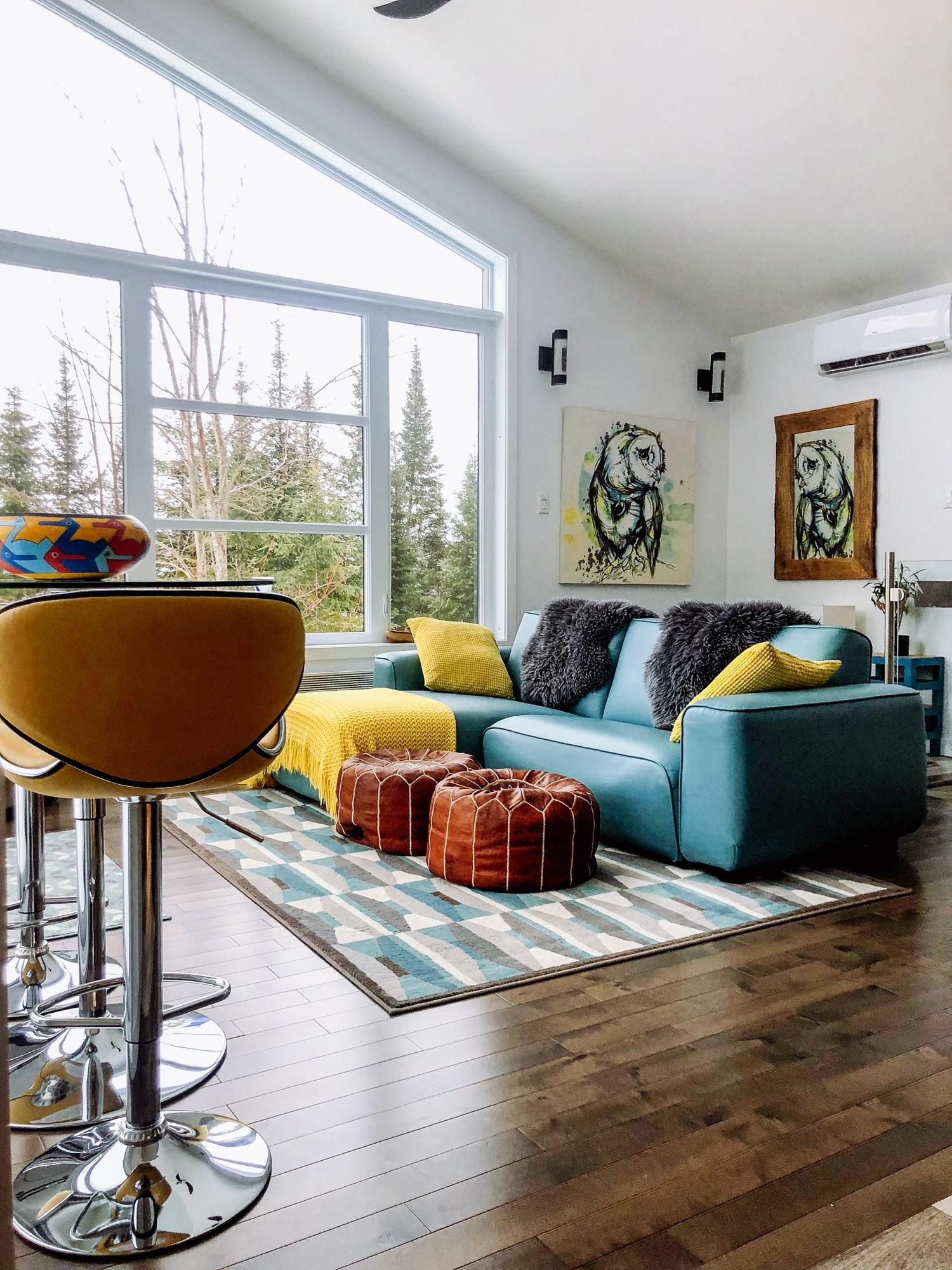 Modern cottage living room with a teal sofa beneath a tall vaulted window looking into pines