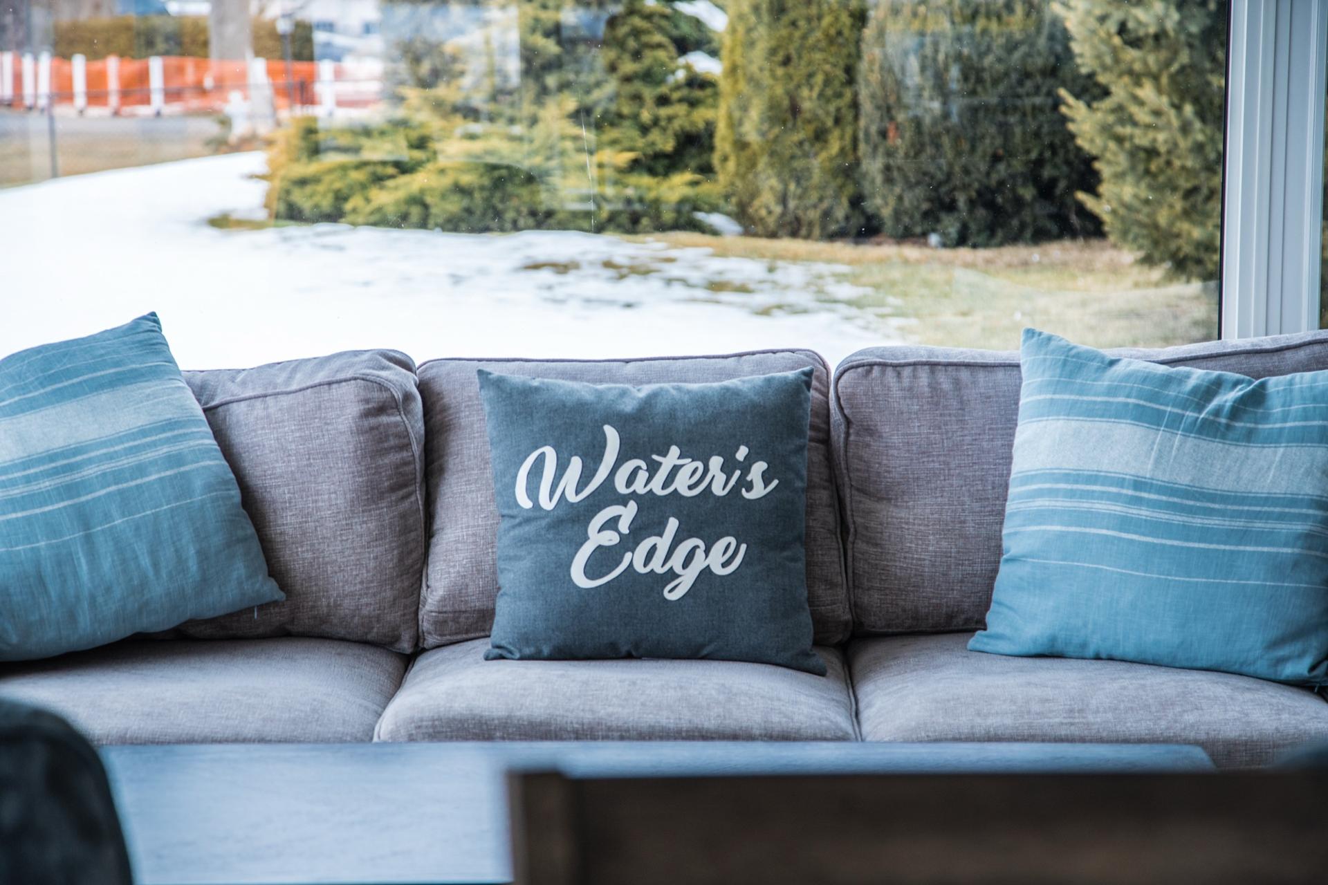 Cushioned sofa by a picture window in a rental cottage, with a cushion reading Water's Edge