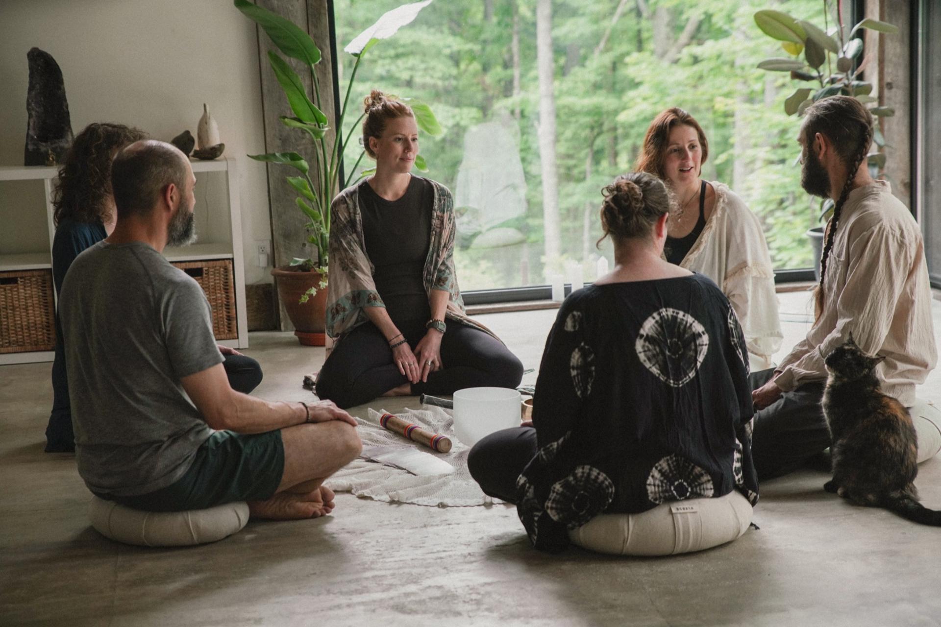 Guests seated in a circle around a crystal bowl, one leading the session, a cat sitting alongside