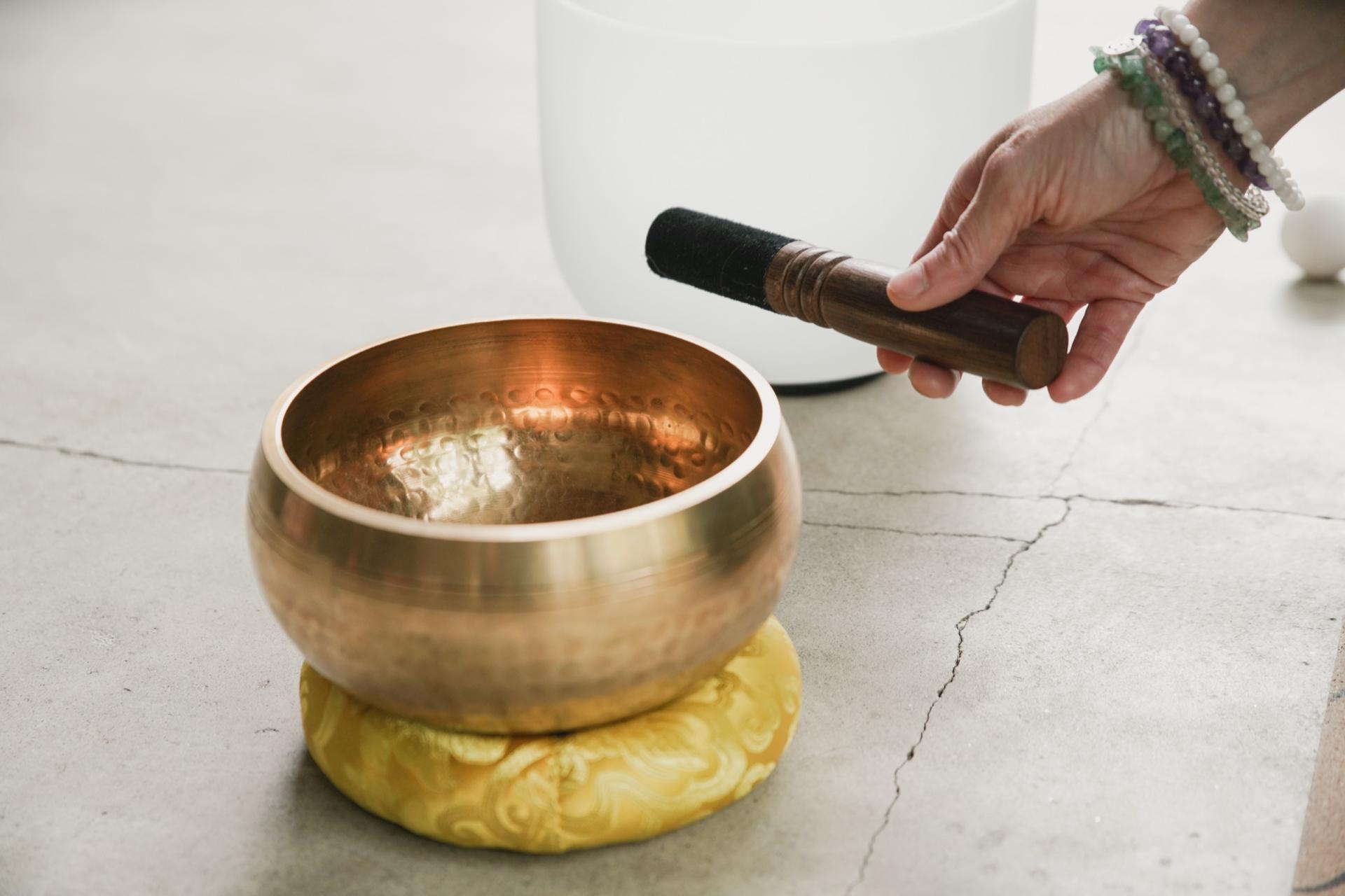 Hand lifting a mallet beside a brass singing bowl resting on a yellow cushion
