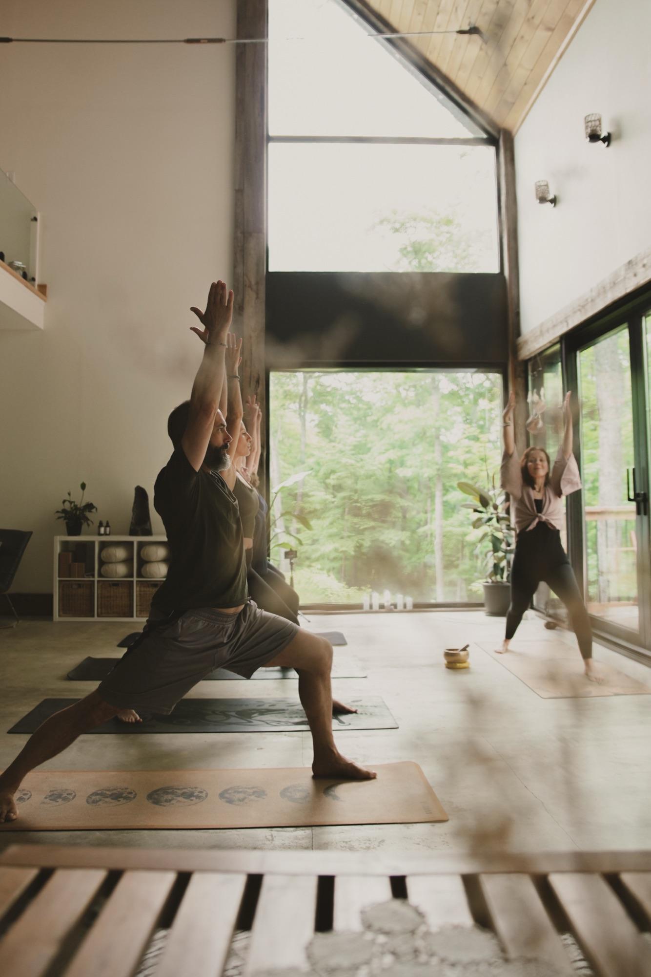 Class in warrior pose with arms raised, the forest visible through tall studio windows