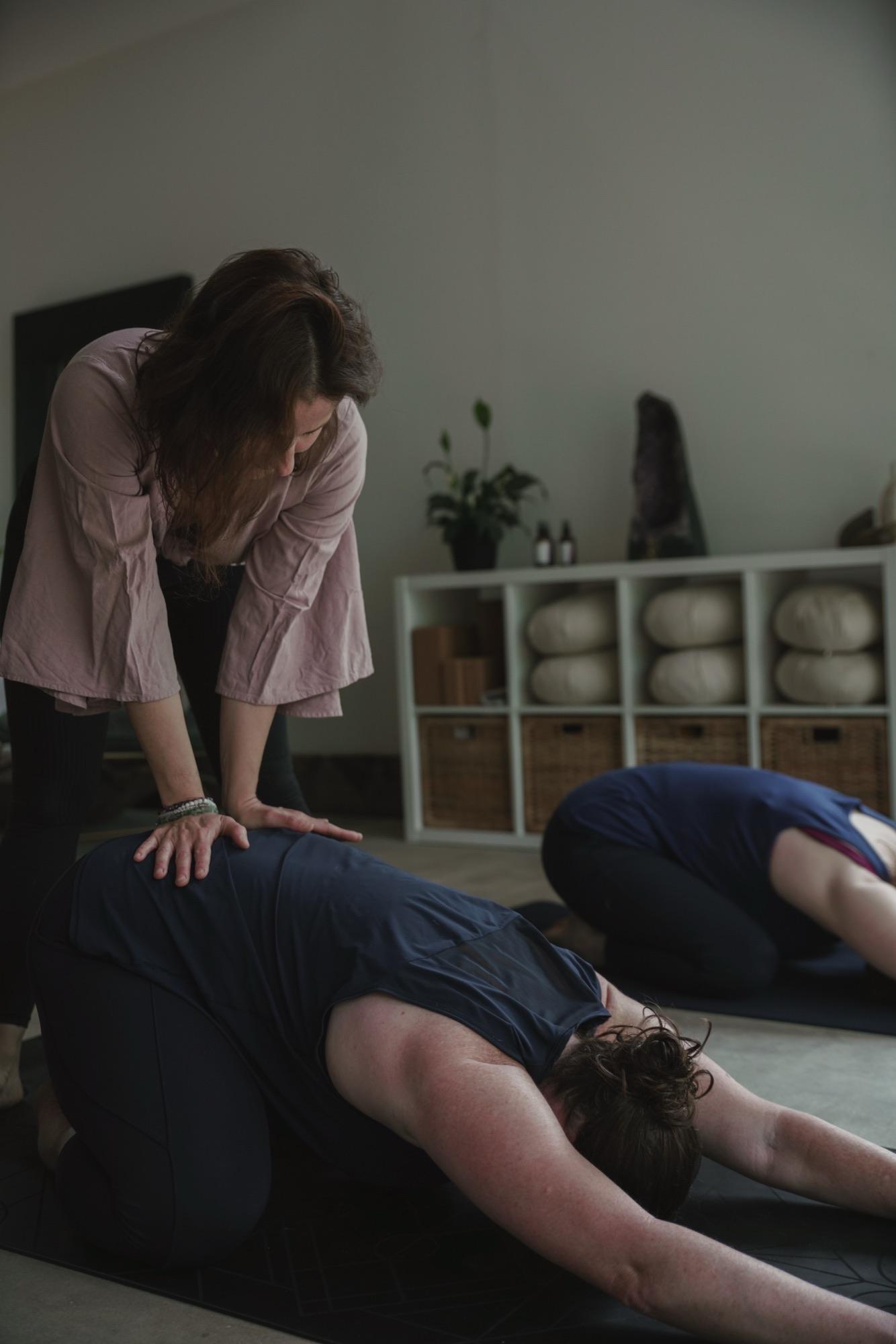 Teacher kneeling to give a hands-on adjustment to a student in child's pose